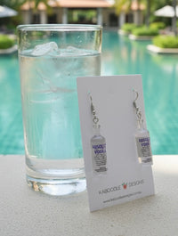 Clear resin vodka bottle drop earrings on a Kaboodle Designs card beside a glass of water