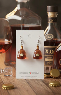 Amber resin XO brandy bottle drop earrings on Kaboodle Designs card, shown against bottles and a glass of cognac