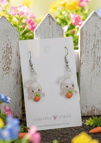 White resin bunny drop earrings with orange carrots on Kaboodle Designs card, garden fence background