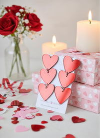 Coral-pink toned heart drop earrings on a card, styled with candles and a red rose bouquet