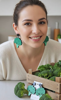 Woman wearing green glitter broccoli dangle earrings, smiling in a kitchen with fresh broccoli nearby
