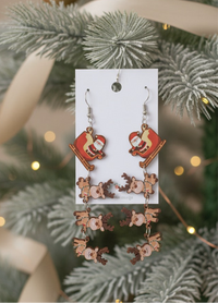 Acrylic Santa sleigh and reindeer drop earrings hanging on card against a frosted Christmas tree