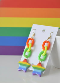 Side view of rainbow chain and star drop earrings displayed against a rainbow striped backdrop