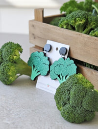 Close-up of broccoli drop earrings on card, propped against a wooden crate filled with fresh broccoli