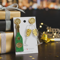 Champagne bottle and toasting glasses earrings displayed on card beside wrapped gifts and fairy lights