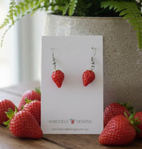 Pair of red resin strawberry drop earrings on a Kaboodle Designs card, surrounded by real strawberries