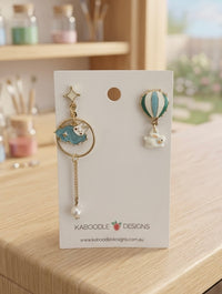 Mismatched earring pair on card: star stud with blue rabbit hoop and pearl drop beside a green hot air balloon with white