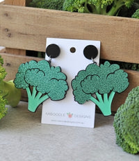 Glittery green acrylic broccoli drop earrings on a black stud, displayed on a Kaboodle Designs card against a wooden crate