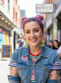 Woman with pink and purple hair wearing rainbow star chain earrings, smiling on a city street
