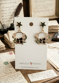 Gold star stud earrings with black enamel cats in a floral wreath, shown on card