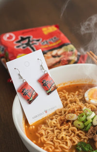 Close-up of Shin Ramyun resin earrings on card with ramen noodle packet in background