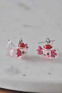 Pair of pink resin axolotl earrings wearing red Santa hats, side view on marble
