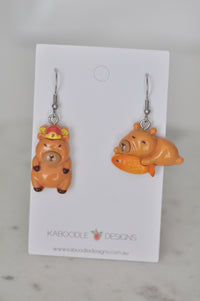 Two mismatched resin capybara earrings on a Kaboodle Designs card, one crowned, one holding a fish