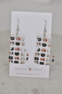 Two cat-shaped perspex drop earrings covered in tiny black, calico and cream kitten face prints on a Kaboodle Designs card