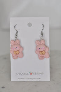 Pair of pink resin bunny rabbit drop earrings with golden bows on white display card