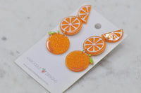 Close-up angled view of glittery acrylic orange earrings with green leaf detail