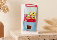 Glossy claw machine brooch featuring colourful googly-eyed monster toys inside a red and blue arcade cabinet design.