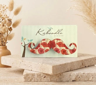 Moustache-shaped brooch featuring a red poppy print design, displayed on a Kaboodle branded card.