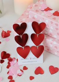 Triple red heart acrylic drop earrings on a Kaboodle Designs card, surrounded by heart confetti and pink gift boxes