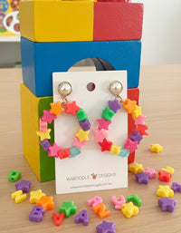 Rainbow star bead hoop earrings on card, displayed against colourful toy blocks with loose star beads