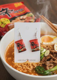 Resin Shin Ramyun packet drop earrings on card, styled beside a steaming bowl of ramen