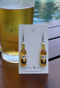 Resin beer bottle drop earrings on Kaboodle Designs card, propped against a glass of beer