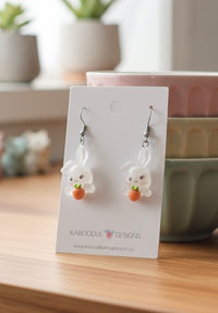 Bunny and carrot resin earrings on card standing beside pastel bowls on wooden bench