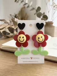 Red smiley face flower stud earrings with black heart top and green leaf stem, on card