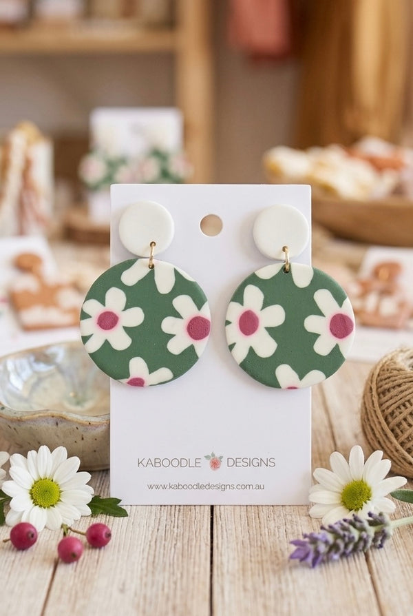 Green clay drop earrings with white daisy print and pink centres on card packaging