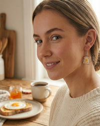 Close-up of a woman wearing a single egg-on-toast earring, showing detail of the fried egg and toast slice