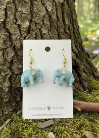Two blue-grey resin elephant drop earrings with gold hooks on a Kaboodle Designs card, propped against a tree trunk
