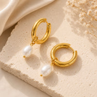 Pair of gold huggie hoops with oval faux pearl drops on stone surface