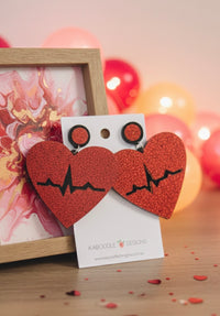 Red glitter heart drop earrings with black heartbeat line, shown on card with balloons and lights behind