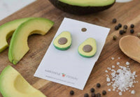 Avocado earrings displayed on wooden board surrounded by sliced avocado and pepper