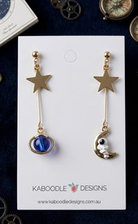 Gold-tone star and blue orb drop earring paired with astronaut on crescent moon earring on white card