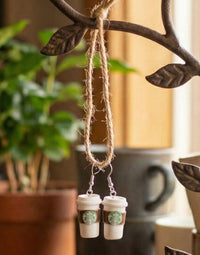Pair of coffee cup earrings hanging from twine against a green plant and mug
