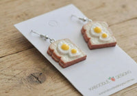 Close-up view of egg-on-toast drop earrings on cardback showing texture of toast crust and glossy egg yolks