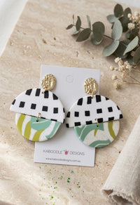 Two-tier clay drop earrings with gold stud, black-and-white checkerboard half-moon, and green swirl round base, shown on card