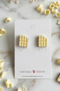White chocolate bar stud earrings with bitten corner detail, flat lay on marble with chocolate curls