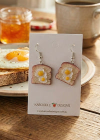 Two resin egg-on-toast drop earrings on a white Kaboodle Designs card, styled with coffee and jam on a wooden table