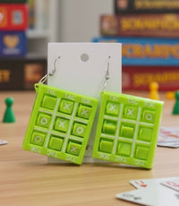 Green plastic tic tac toe drop earrings hanging on a display card, board games in background