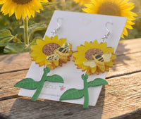 Close-up front view of layered acrylic sunflower earrings with bee detail and dangling stem