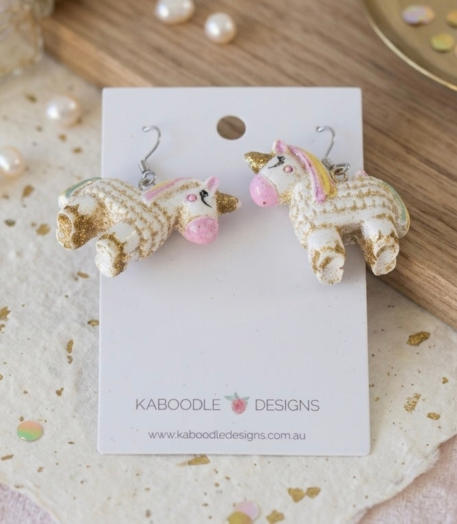 Unicorn pinata dangle earrings laid flat on Kaboodle Designs card among pearls and gold confetti