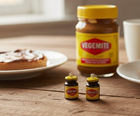 Two Vegemite jar earrings standing upright on a wooden table beside a full-size Vegemite jar and toast