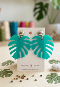 Teal perspex monstera leaf drop earrings with silver ball studs on a Kaboodle Designs card