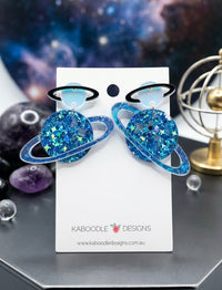Pair of blue glitter Saturn planet drop earrings with black rings on Kaboodle Designs card