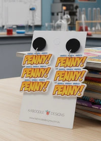 Acrylic dangle earrings with triple 'PENNY!' text in yellow and red, shown on display card