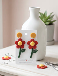 Red acrylic flower drop earrings with mustard stud top on Kaboodle Designs card, styled with a white vase
