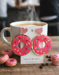 Pink iced donut drop earrings with rainbow sprinkles on a Kaboodle Designs card, propped against a coffee cup