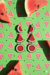 Watermelon slice earrings laid flat on green watermelon-print background beside real watermelon wedges